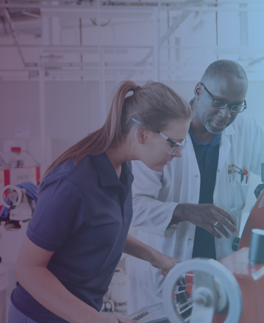 Older man helps younger woman understand a piece of machinery in a lab